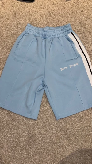 Shorts - shorts i ljusblått med vit och svart rand längs sidorna. Elastisk midja med snörning och två fickor framtill. Tillverkade i ett mjukt, sportigt polyestermaterial. Loggan är tryckt på vänster ben i vitt.