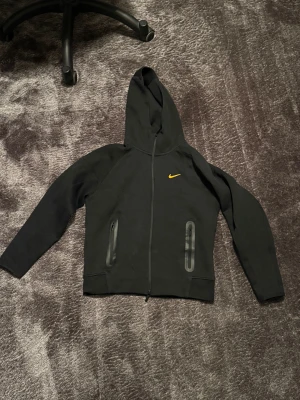  Nike NOCTA zip-Hoodie  - Svart hoodie från Nike NOCTA med gul swoosh på bröstet. Tröjan har hel dragkedja, två fickor med dragkedja och stor huva. Materialet är mjuk bomull och polyester, perfekt för chill eller träning. Snygg och clean design som sticker ut. 