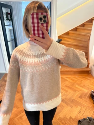 Beige stickad tröja med mönster - Supermysig beige stickad tröja med vitt och ljusrosa mönster runt halsen och vid muddar. Rund hals och långärmad, perfekt för kyliga dagar. Tröjan har en loose passform och ribbade muddar för extra komfort.