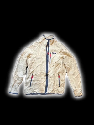 Beige teddyjacka från Patagonia - Säljer en beige teddyjacka från Patagonia med marinblå detaljer och dragkedjor. Jackan har tre fickor med dragkedja och coola röda zip-pullers. Insidan har ett blått meshfoder på ena sidan. Perfekt för dig som gillar chill och varm stil.