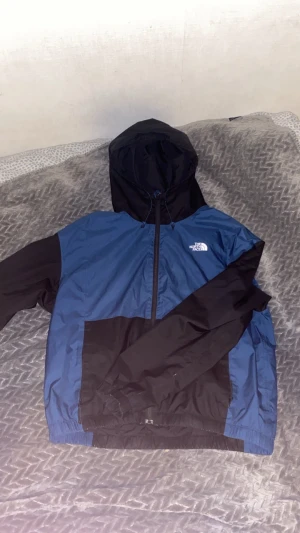 Blå och svart vindjacka från The North Face - Snygg blå och svart vindjacka från The North Face med huva och dragkedja framtill. Jackan har elastiska muddar, logga på bröstet och är tillverkad i lätt polyester. Perfekt för dig som gillar streetstyle och vill ha en clean look.