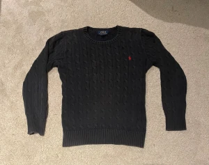 Mörkblå tröja från Polo Ralph Lauren - Snygg mörkgrå stickad tröja från Polo Ralph Lauren med klassiskt kabelstickat mönster och röd broderad logga på bröstet. Perfekt för dig som gillar stilrena och tidlösa plagg.