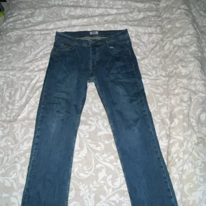 Tommy Hilfiger Jeans blåa - Säljer ett par klassiska blå jeans från Tommy Hilfiger Jeans med snygga kontrastsömmar och ikonisk logga på bakfickan. Modellen har normal passform och raka ben, perfekt för dig som gillar en tidlös look. Materialet är slitstarkt denim och midjan är normal. Kan diskutera om priset!