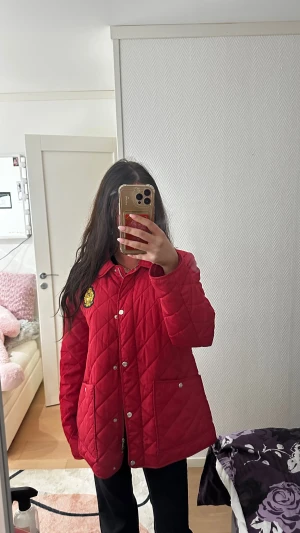 Röd quiltad jacka från Ralph Lauren - Säljer en snygg röd quiltad jacka från Ralph Lauren med guldigt broderat emblem på bröstet. Jackan har krage, tryckknappar framtill och två stora fickor. Materialet är glansig polyester och passformen är normal. Perfekt för dig som vill sticka ut med stil.