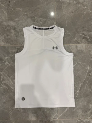 Vit linnetopp från Under Armour - Säljer en vit linnetopp från Under Armour med ventilerande meshmaterial och logga i silver på bröstet. Modellen är ärmlös och har rund halsringning med svart text på insidan. Perfekt för dig som gillar sportig och clean stil.