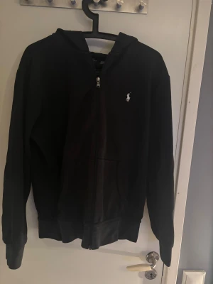 Svart hoodie från Polo Ralph Lauren - Svart hoodie från Polo Ralph Lauren med klassisk broderad logga på bröstet. Tröjan har dragkedja framtill, huva och två fickor. Tillverkad i mjuk bomull och har långärmad passform. Perfekt för chill dagar och enkel att matcha med jeans eller joggers.