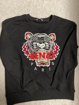 Svart Kenzo sweatshirt med tiger - Svart sweatshirt från Kenzo med ikonisk broderad tiger och logga i rött, vitt och grönt på bröstet. Tröjan har rund hals, ribbade muddar och är tillverkad i mjuk bomull. Perfekt för dig som gillar statement-plagg och streetstyle.