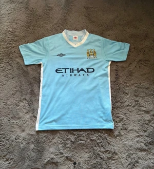 Manchester City Nasri 19 matchtröja - Ljusblå Manchester City fotbollströja från Umbro med Nasri och nummer 19 på ryggen. Tröjan har klubbmärke och Etihad Airways-tryck på bröstet, vita detaljer längs sidorna och klassisk V-ringning. Perfekt för dig som vill visa ditt City-stöd.