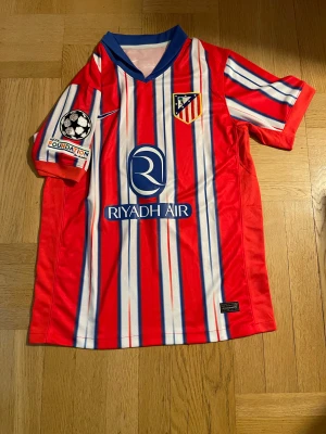 Atlético Madrid matchtröja Nike röd/blå - Säljer en officiell Atlético Madrid fotbollströja från Nike med vertikala röd, vit och blå ränder, klubbmärke på bröstet och UEFA Foundation-märke på ärmen. Tröjan har blå krage, korta ärmar och är tillverkad i lätt polyester.