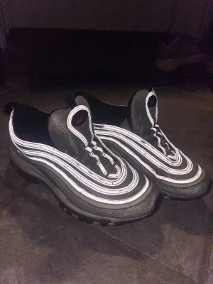 Nike Air Max 97 Silver Bullet sneakers - Nike Air Max 97 Silver Bullet sneakers med ikoniska vågiga linjer i silver och reflekterande detaljer. Röd swoosh på sidan och tungan, svart innersula och platt sula med synlig Air-enhet. Materialet är syntet och skinn, med snörning och rund tå. Perfekt för dig som gillar klassiska och sportiga sneakers.