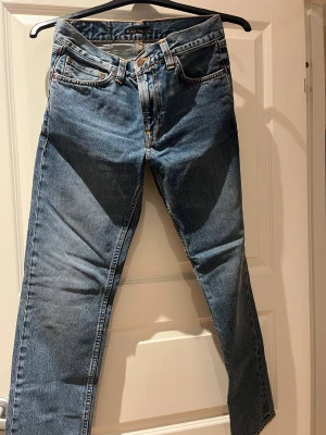 Blå jeans från Nudie Jeans, W28 L30 - Säljer ett par klassiska blå jeans från Nudie Jeans med orange sömmar och snygg broderad detalj på bakfickorna. Modellen har rak passform och normal midja. Tillverkade i slitstarkt bomullsjeans-tyg. Perfekta för dig som gillar tidlös stil.