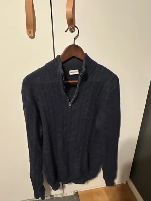 Mörkblå stickad tröja John Henric - Snygg mörkblå stickad tröja från John Henric med kabelstickat mönster och half zip-dragkedja vid halsen. Tröjan har ribbad krage och långa ärmar, perfekt för dig som gillar klassisk och stilren look. Storlek M.