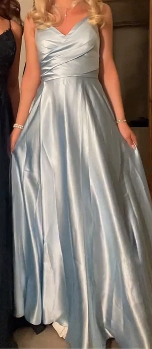 Ljusblå balklänning med snörning  - Säljer en jättefin ljusblå långklänning i glansigt satinliknande material. Klänningen har omlottliv, v-ringning och smala axelband med snygg snörning i ryggen. Kjolen är vid och faller elegant. Perfekt för bal eller student! Använt en gång. Jag är 162cm och jag har klackar på bilden. Man kan styra mycket hur den ska sitta I midjan. Betalade 2800 för den, pris kan diskuteras 