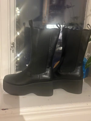 Svarta plattform boots från NLY Shoes - Säljer ett par  helt nya svarta boots med rejäl plattformssula från NLY Shoes. Skorna har rund tå, elastiska paneler på sidorna och är tillverkade i syntetmaterial med skinnlook. Perfekt för dig som vill ha extra höjd och en edgy stil . Storlek 37 