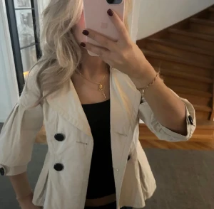 Beige kavajjacka - Snygg beige kavajjacka med bred krage och stora svarta knappar framtill. Jackan har trekvartsärmar med knappdetalj vid ärmslut och en loose passform. Perfekt att slänga över en topp för en cool och stilren look. Mer som en S än M. Har en liten fläck men syns inte vid användning