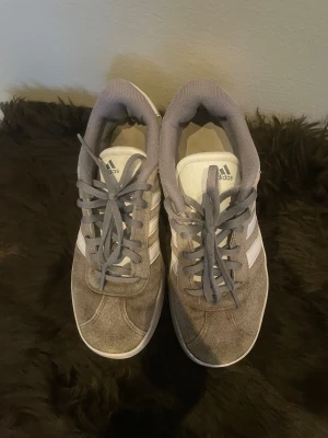 Gråa Adidas sneakers i mocka - Snygga gråa Adidas sneakers med klassiska vita ränder på sidorna. Ovandelen är i mjuk mocka och skorna har grå snörning samt vit sula. Perfekta för en chill och sportig stil. Låg modell med rund tå och platt sula.