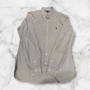 Rutig skjorta från Ralph Lauren - Snygg rutig skjorta från Ralph Lauren i beige och blå toner. Klassisk button-down krage, långärmad och med knappar framtill. Broderad logga på bröstet. Skjortan är gjord i mjuk bomull och har en normal passform som funkar till många stilar.