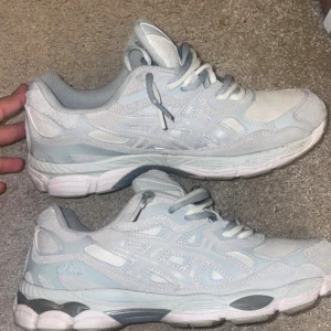 Asics gel nyc - Dubbellacade. Säljer ett par fräscha Asics sneakers i vitt och ljusblått. Skorna har en sportig vibe med mesh och syntetpaneler, samt klassiska Asics-detaljer på sidorna. De har snörning och en bekväm, vadderad insida. Perfekta för dig som gillar stilrena och bekväma sneakers.