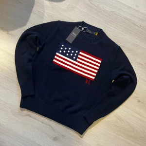Mörkblå stickad tröja Polo Ralph Lauren - Mörkblå stickad tröja från Polo Ralph Lauren med amerikansk flagga i vitt och rött på bröstet samt RL-broderi. Rund halsringning och ribbade muddar vid ärmslut och nederkant. Perfekt för dig som gillar klassisk och stilren design. Är i M men sitter som S