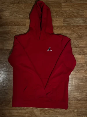 Röd Jordan hoodie med logga - Snygg röd hoodie från Jordan med klassisk Jumpman-logga broderad på bröstet. Tröjan har huva, långa ärmar och en stor magficka. Mjuk insida i bomullsmix som gör den skön att bära. Perfekt för dig som gillar streetwear och sportig stil.