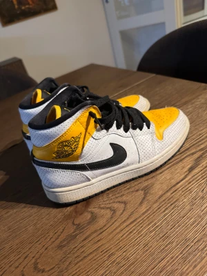 Nike Air Jordan 1 High vit/gul - Nike Air Jordan 1 High sneakers med perforerad vit ovandel, gula detaljer på tå och skaft, samt svart swoosh och snörning. Klassisk Jordan-logga på sidan och svart plös med Jumpman. Skorna har en rund tå och platt sula, tillverkade i skinn.