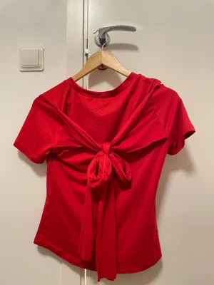 Röd kortärmad topp med knyt fram - Säljer en röd topp med korta ärmar och rund hals. Framsidan har en snygg knyt-detalj som ger en cool twist. Materialet känns mjukt och stretchigt, perfekt för dig som gillar att sticka ut med färg. Passformen är normal och toppen är enkel men ändå unik.