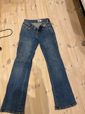 Blå bootcut jeans från 157 Paris Stitch - Snygga blå jeans från 157 Paris Stitch med klassisk bootcut passform. Jeansen har kontrastsömmar i gult, fem fickor och knappstängning framtill. Materialet är robust denim som ger en cool vintagekänsla. Perfekt för dig som gillar retrostil och vill sticka ut.