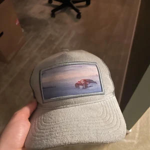 Grå keps från Revenite med bilmotiv - Snygg grå keps från Revenite med ett broderat motiv av en bil mot solnedgång på framsidan. Kepsen har justerbar snapback i plast och mesh på insidan för extra ventilation. Perfekt för dig som gillar coola accessoarer med biltema.