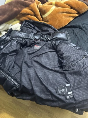 Svart pufferjacka från Prada - Säljer en svart pufferjacka från Prada med glansig finish och huva. Jackan har dragkedja, tryckknappar och två fickor framtill. Insidan är mönstrad med Prada-loggor och jackan har snörning vid huvan för extra stilpoäng. Perfekt för kalla dagar.