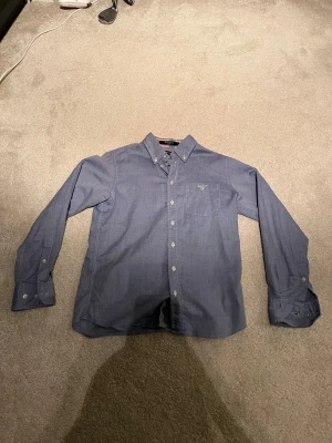 Blå Oxfordskjorta från GANT 146-152  - Snygg blå Oxfordskjorta från GANT i storlek 146-152. Skjortan har klassisk button-down krage, bröstficka med broderad logga och vita knappar. Materialet är mjuk bomull och passformen är normal. Perfekt för dig som gillar stilrena och tidlösa plagg. Bara använd ett par gånger 