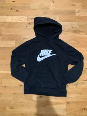 Svart Nike hoodie i bra skick - Fin Svart hoodie från Nike i storlek 158-170. Den är väldigt bra skick och har inga skador. Den har en stor framficka. Materialet inuti är mjukt och skönt och själva tröjan är inte så tjock eller så tunn. Hör av dig vid funderingar!