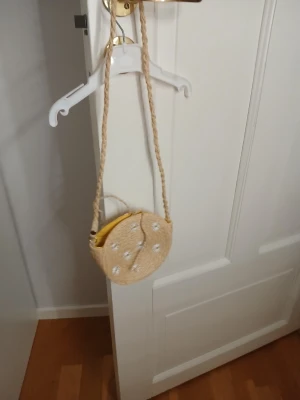 Rund axelväska i flätad strå - Säljer en rund axelväska i naturfärgad flätad strå med långt axelband. Väskan har broderade vita blommor på framsidan och gul insida. Perfekt för dig som vill ha en chill och somrig look.