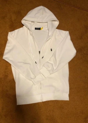 Vit hoodie från Polo Ralph Lauren - De står att storleken är l men känns som en M och den är lite smutsig Vit hoodie från Polo Ralph Lauren med dragkedja och huva. Klassisk design med den lilla broderade loggan på bröstet och ribbade muddar. Perfekt för chill dagar och enkel att matcha med jeans eller joggers.