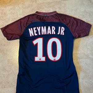  Neymar Jr PSG fotbollströja  - Paris Saint-Germain fotbollströja med Neymar Jr och nummer 10 på ryggen. Tröjan är marinblå med vinröda korta ärmar, har broderat PSG-emblem och Nike-logga samt Fly Emirates-tryck på bröstet. Tillverkad i lätt polyester med Dri-FIT-teknologi.