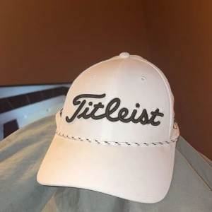 Vit Titleist keps med broderad logga - Snygg vit keps från Titleist med stor broderad logga i svart framtill. Klassisk böjd skärm och dekorativ söm längs kanten. Perfekt för golf eller som cool accessoar till vardags. Justerbar baktill för bästa passform.