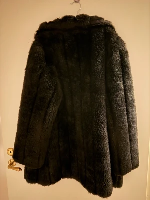 Svart pälsjacka med klassisk look - Säljer en svart pälsjacka med mjuk och fluffig känsla. Jackan har en rak passform, lång ärm och en enkel krage. Perfekt för dig som vill ha en stilren och varm jacka med lyxig vibe. Färgen är djup svart och materialet är pälsimitation.