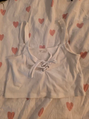 Vit ribbad linnetopp från Stories - Supersöt vit ribbad linnetopp från Stories i storlek S. Toppen har breda axelband och snygg snörning framtill med metallöglor. Croppad modell som är perfekt till jeans eller kjol. Materialet är mjukt och stretchigt för en skön passform.