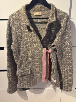 Grå mönstrad kofta Odd Molly - Superfin kofta från Odd Molly i grått och vitt med snyggt stickat mönster. Jackan har lång ärm, krage, rosa rosetter och knytband framtill. Broderad logga på ryggen och fickdetaljer. Perfekt för dig som gillar unika detaljer och mysig stil.