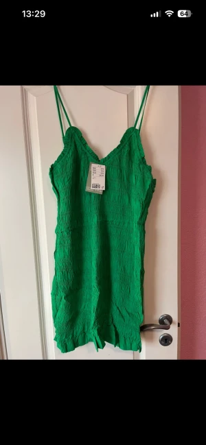 Grön smockad kortklänning med volang från H&M storlek L - Superfin grön kortklänning från H&M med smockad struktur, tunna axelband och volangdetalj längst ner. Perfekt för varma dagar och enkel att styla med sneakers eller sandaler. Klänningen har en v-ringning och är i ett lätt material.