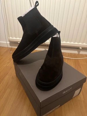 GANT Vinterskor Mocka – Helt Nya - Säljer ett par mörkbruna herrskor från GANT i mocka. Skorna är helt nya och endast provade kort inomhus, aldrig använda utomhus. De kommer i originalkartong och är i perfekt nyskick. Detta är ett stilrent och kvalitativt par som fungerar utmärkt både till vardag och mer uppklädda tillfällen. Ordinarie pris ligger runt 2000kr, men säljs då de tyvärr inte passade mig i storlek.