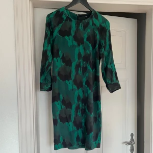 Kort klänning i gröna toner - Klänning från Just Female i mjukt, lätt stretchigt syntetmaterial med underbart fall. Modell som kan stylas med skärp eller loose fit med vackert veckad skräddad söm baktill. Passar både fest och vardag. Grön botten med mörkgrå - svart/jordig nyans i mönstret. Diskret glans i tyget och osynlig dragkedja bak.  Passar strl S/36-38