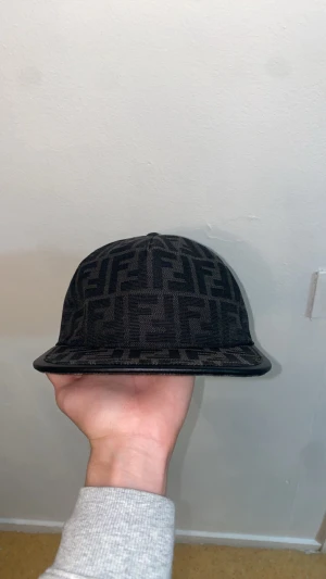 Svart Fendi keps med FF-mönster - Säljer en svart Fendi keps med ikoniskt FF-mönster över hela kepsen. Kepsen har rak skärm och justerbar rem baktill med Fendi-logga. Materialet är mjukt och känns lyxigt, perfekt för dig som gillar designer och streetwear.