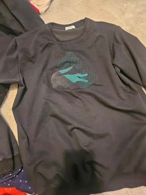 Svart sweatshirt  - Svart sweatshirt med rund halsringning och stort tryck av den klassiska krokodilen i turkos och grått framtill. Tröjan har långa ärmar och är tillverkad i mjuk bomull, perfekt för en avslappnad stil.