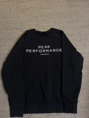 Svart Peak Performance sweatshirt M - Svart sweatshirt från Peak Performance i storlek M junior. Tröjan har rund halsringning, långa ärmar och vit logga tryckt framtill. Klassisk och stilren modell i mjukt bomullsmaterial, perfekt för en avslappnad look.