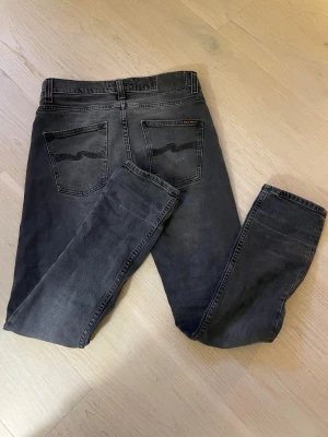 Svarta jeans från Nudie Jeans - Säljer ett par svarta  jeans från Nudie Jeans i storlek 29/30. Jeansen har klassisk femficksdesign, svarta sömmar och Nudie Jeans logga på bakfickan. Materialet är mjukt denim och passformen är slim.