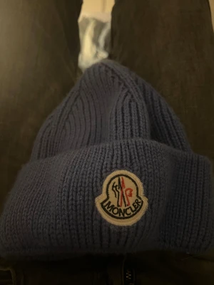 Blå stickad Moncler mössa - Snygg blå ribbstickad mössa från Moncler med klassisk logotyp framtill. Mössan har uppvikt kant och är gjord i mjukt stickat material som håller dig varm. Perfekt för dig som vill ha en clean och stilren look med lite lyxig känsla.