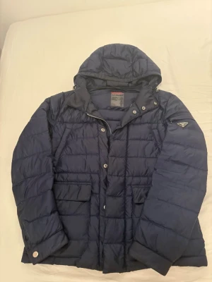 Mörkblå dunjacka från Prada - Säljer en mörkblå dunjacka från Prada med huva och quiltad design. Jackan har flera fickor med lock, dragkedja och tryckknappar framtill samt ikonisk Prada-logga på ärmen. Yttermaterialet är polyester och fodret består av dun och fjäder. Två knappar har ramlat av från jackan annars inga andra defekter. Pris kan diskuteras ifall du är intresserad. Jackan är storlek 48 och borde passa om man är 182-190