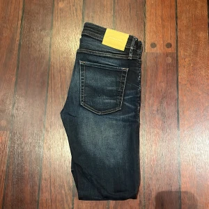 Skinny jeans Liam mörkblå 30/32 - Mörkblå skinny jeans från Jack & Jones, modell Liam. Snygg tvätt med slitningar och klassiska fem fickor. Jeansen har smal passform och är tillverkade i stretchigt denim för extra komfort. Perfekta för dig som gillar en tajt look.