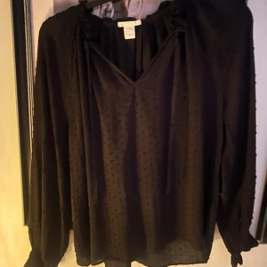 Svart blus med knyt och prickar - Säljer en svart blus med diskret prickigt mönster och v-ringning. Blusen har långa ärmar med resår vid ärmslut och knytband vid halsen. Tunt, lätt material som ger en luftig känsla och snyggt fall.