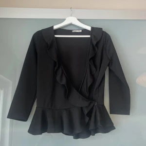 Svart omlottopp med volang från Nelly Trend - Svart omlottopp från med v-ringning. Toppen har volangdetaljer längs ringningen och nertill, samt knäppning i sidan. Snygg och feminin passform som framhäver midjan.